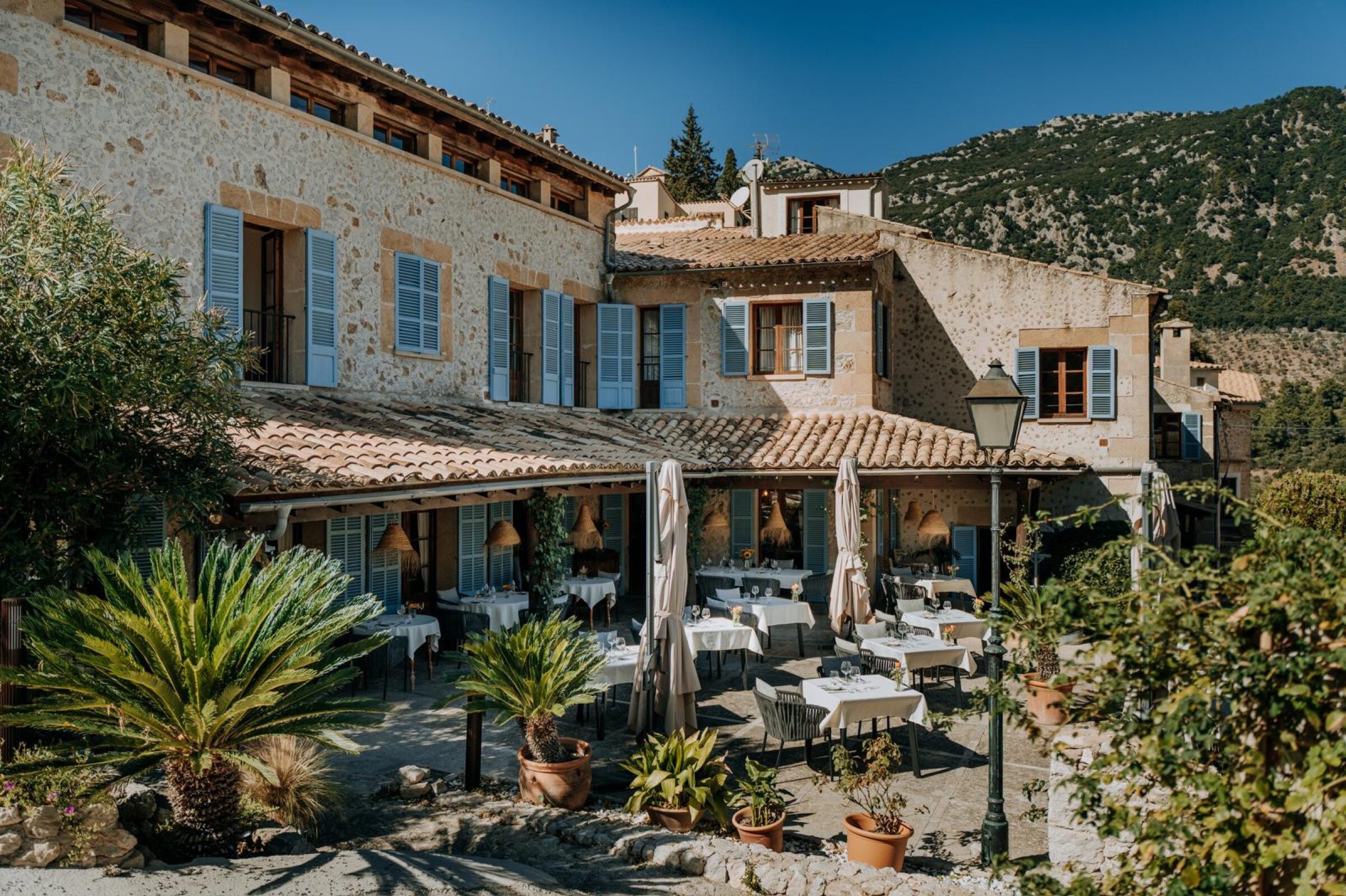The stone façade of Nou Dalt Muntanya with sage-blue shutters and a tiled terrace of white-clothed tables, a wooded Tramuntana ridge rising directly behind it under a deep blue sky