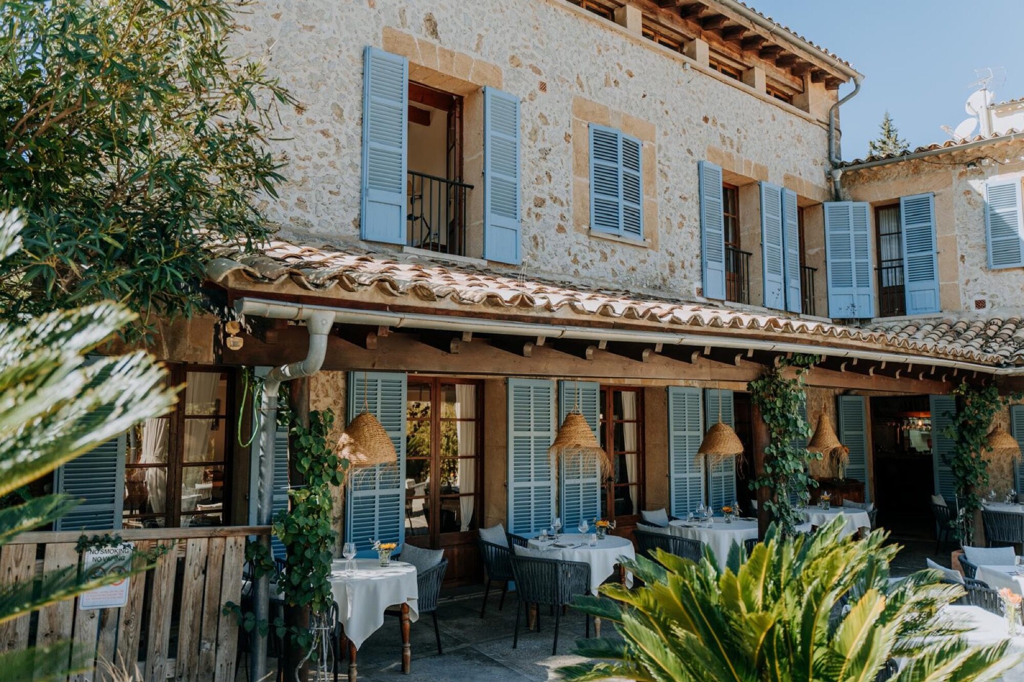 The front of the hotel: a stone façade with blue shutters and a covered terrace of set tables, framed by palms and potted plants