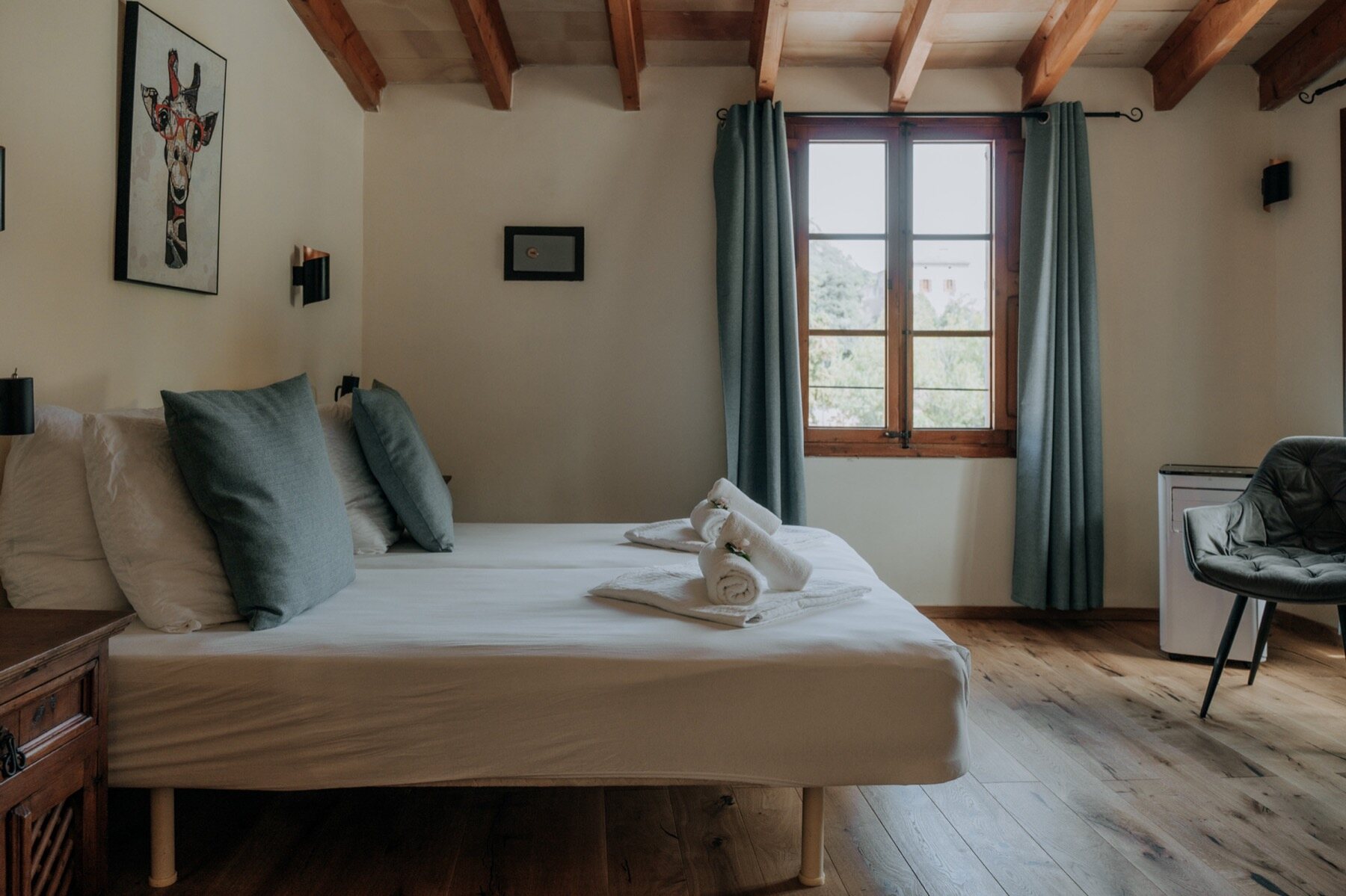A bright double room under exposed beams with sage-blue curtains, oak floor, white linen and a shuttered window onto greenery
