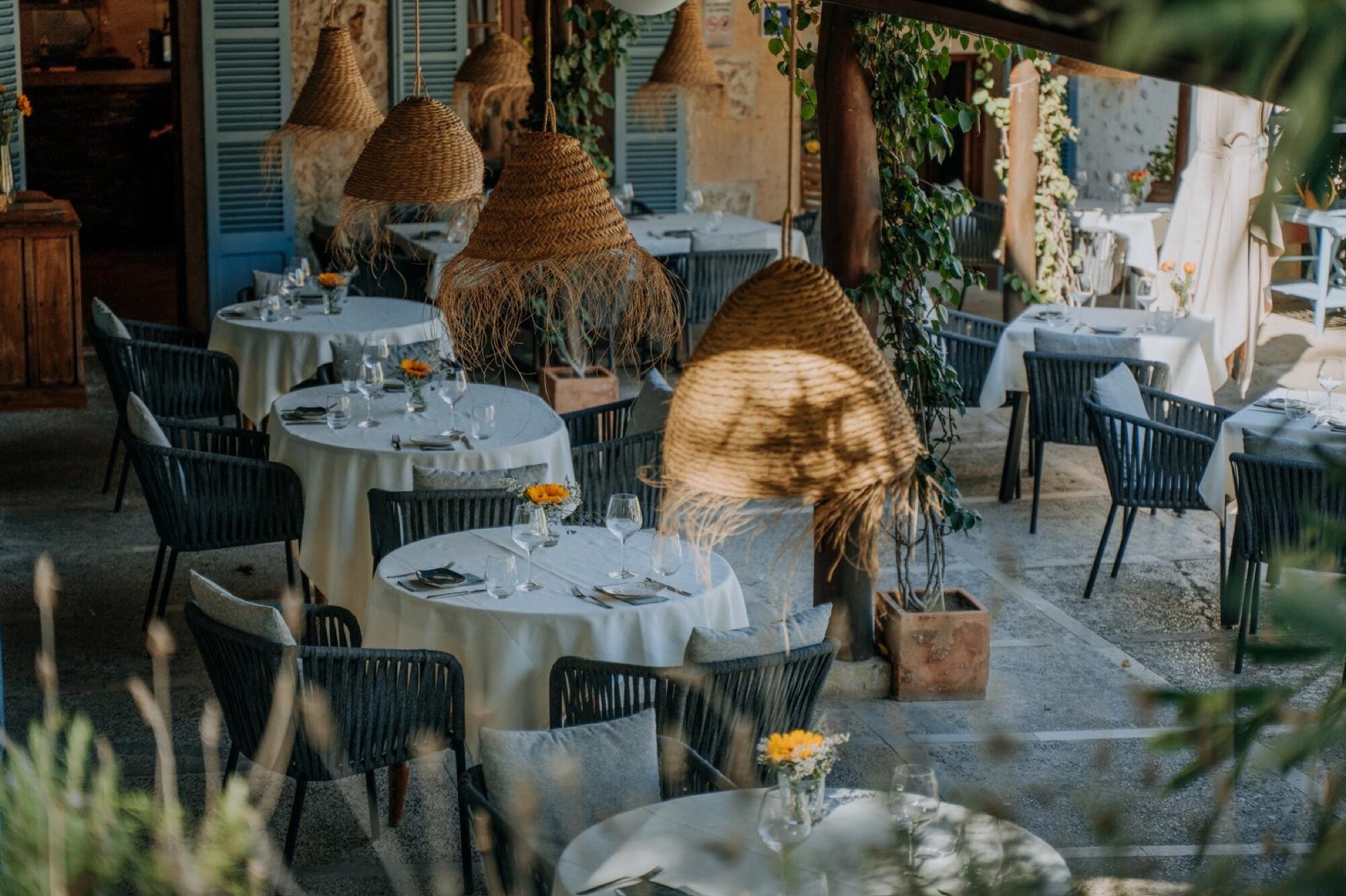 The Es Freu terrace laid for dinner under woven basket pendant lamps, black rattan chairs and white linen, marigold stems on each table