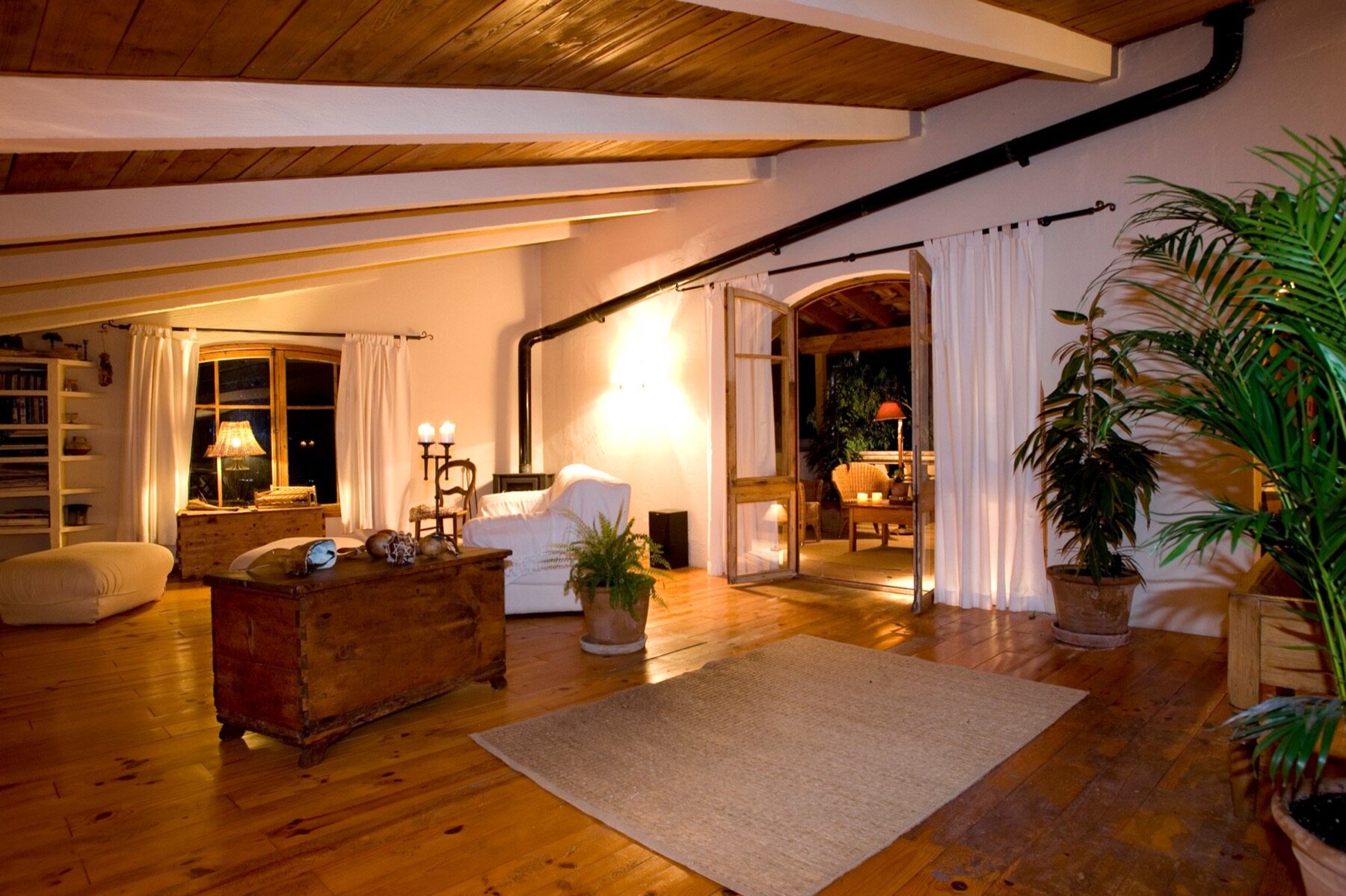 A warm sitting room with exposed timber beams, deep sofas and tiled floors, lit low in the evening